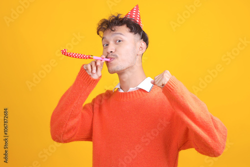 Portrait of Young Asian Man Wear Birthday Cap Blowing Whistle While Celebrating Clencing Fist Isolated on Yellow Background photo