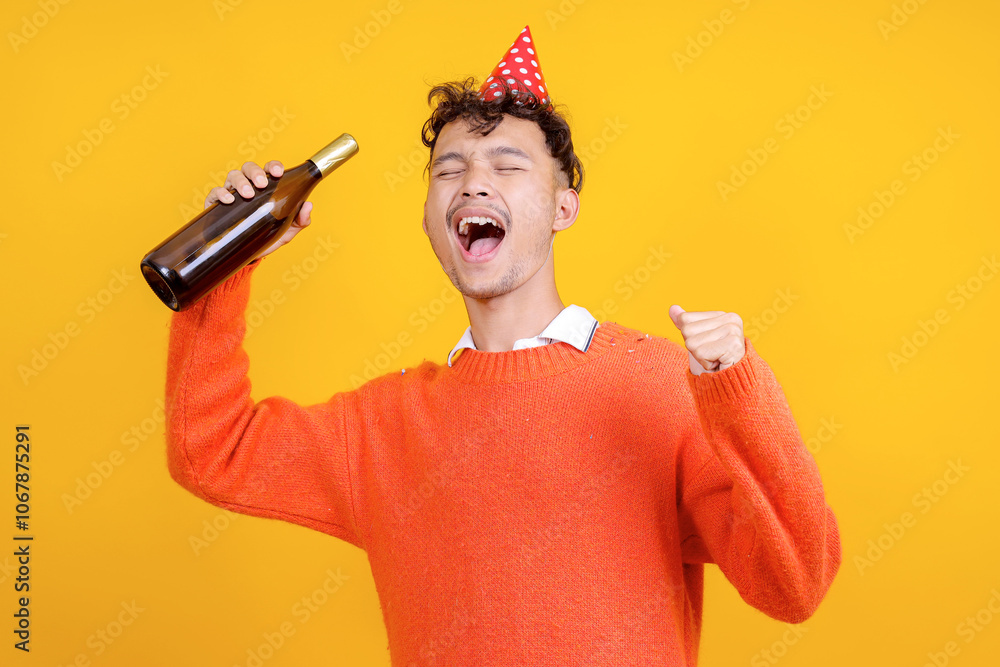 Excited Young Asian Man Clenching Fist Celebrating Victory While Holding Wine Bottle Isolated on Yellow Background Excited Young Asian Man Clenching Fist Celebrating Victory While Holding Wine Bottle Isolated on Yellow Background