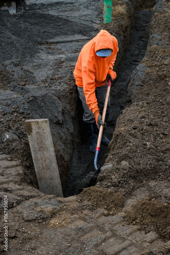 Construction worker in a trench dug out to install underground PVC pipe conduits, wearing an orange safety sweatshirt and working with a shovel 