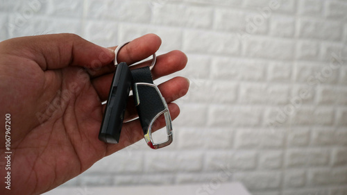 A close-up shot of a hand holding a keychain with a black leather strap and a metal carabiner clip, set against a textured white brick wall background. Ideal for lifestyle and accessory themes.