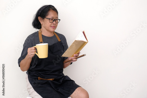 Asian housewife wearing black apron, sitting alone enjoying hot coffee and reading recipe book photo