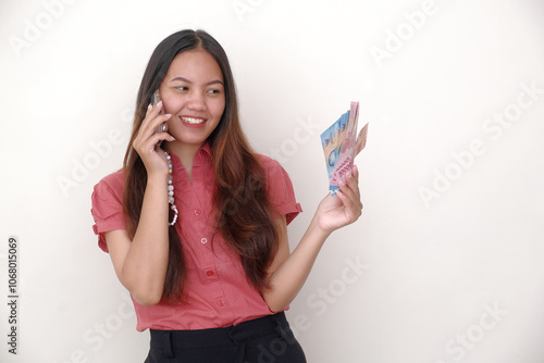 A young Asian girl making a phone call with her smartphone, while other hand holding some cash Rupiah money photo