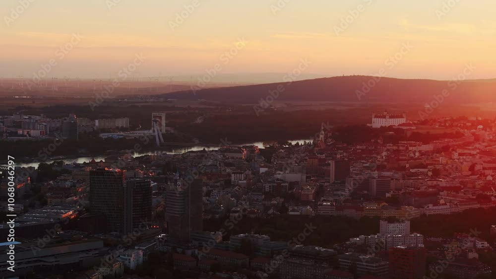 The sun sets over Bratislava, Slovakia, casting an orange glow over urban buildings, the Danube River, and distant UFO Bridge The sun sets over Bratislava, Slovakia, casting an orange glow over urban buildings, the Danube River, and distant UFO Bridge