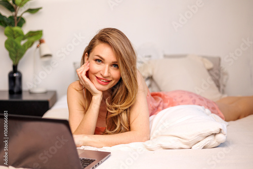 A woman smiles while resting on a bed, using a laptop and enjoying her time in a cozy bedroom