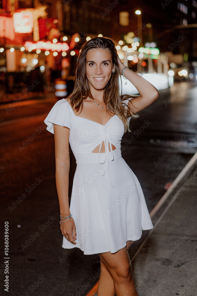 A stylish young woman confidently poses in a flowing white dress against a vibrant and colorful city backdrop at night