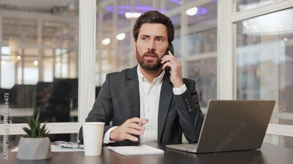 Busy businessman in a suit sits at his desk in modern office, talking on cell phone and gesturing to the laptop. Productive work environment, with businessman focused on his tasks during busy workday. Busy businessman in a suit sits at his desk in modern office, talking on cell phone and gesturing to the laptop. Productive work environment, with businessman focused on his tasks during busy workday.