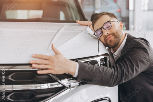 Positive young bearded caucasian man in glasses hugging his new car in dealership. Happy man finally gets long-awaited car, wearing formal suit