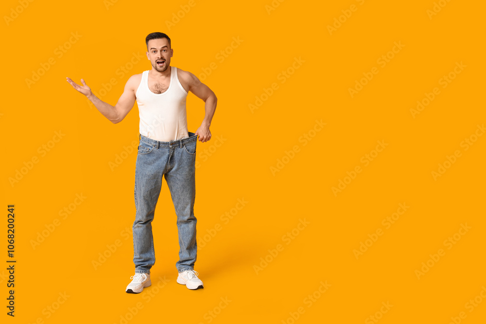 Fototapeta premium Handsome sporty shocked man in loose jeans on yellow background. Weight loss concept