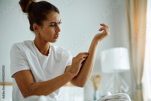 Bild auf Leinwand Young woman looking at her itchy arm skin in bedroom.