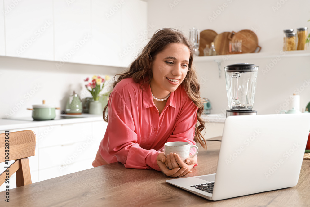Beautiful young woman with coffee cup and laptop on table in kitchen Beautiful young woman with coffee cup and laptop on table in kitchen