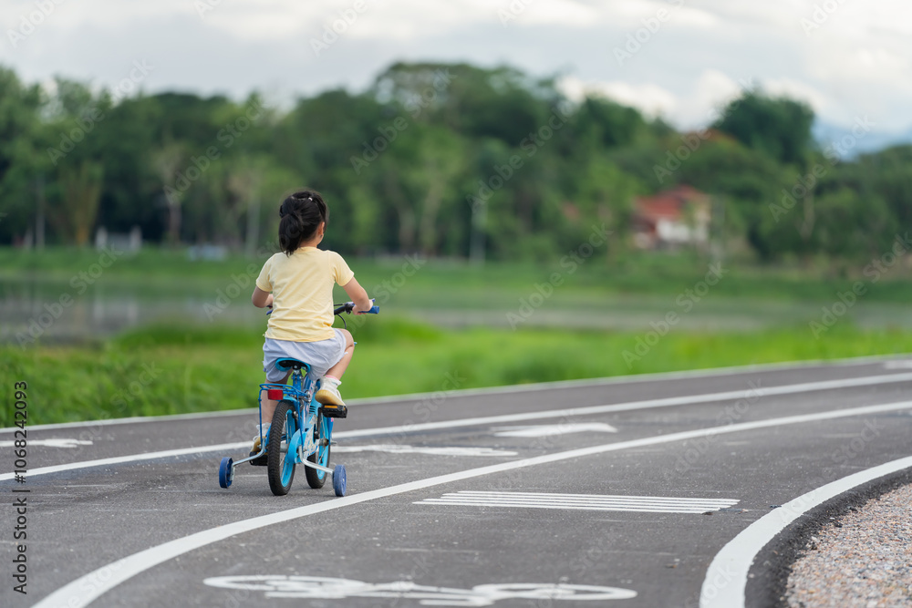 Obraz premium Asian young girl is riding a blue bicycle down a road. The road is lined with trees and there is a house in the background. The scene is peaceful and serene, with the girl enjoying her ride