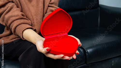 Asian woman sitting on black sofa and holding red heart shaped gift box with gold bow in hand. Gift box open lid, present and gift boxes for festival and celebration.