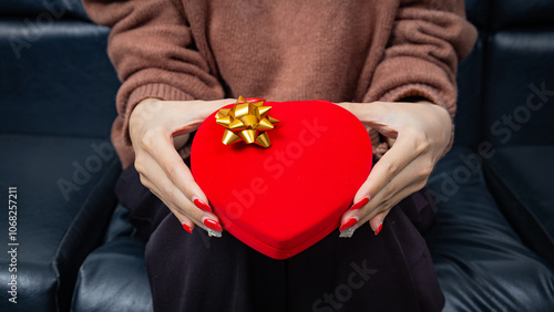 Asian woman sit on black sofa and holding a red heart shaped gift box with a gold ribbon bow in hands. Present and gift boxes for festivals and celebrations.
