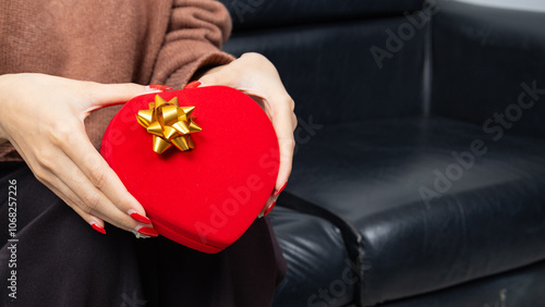 Asian woman wearing a brown shirt sit on black sofa and holding a red heart shaped gift box with a gold ribbon bow in hands. Red gift box. Presents and gift boxes for festivals and celebrations.