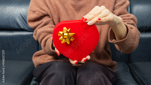 Asian woman wearing a brown shirt sit on black sofa and holding a red heart shaped gift box with a gold ribbon bow in hands. Red gift box. Presents and gift boxes for festivals and celebrations.