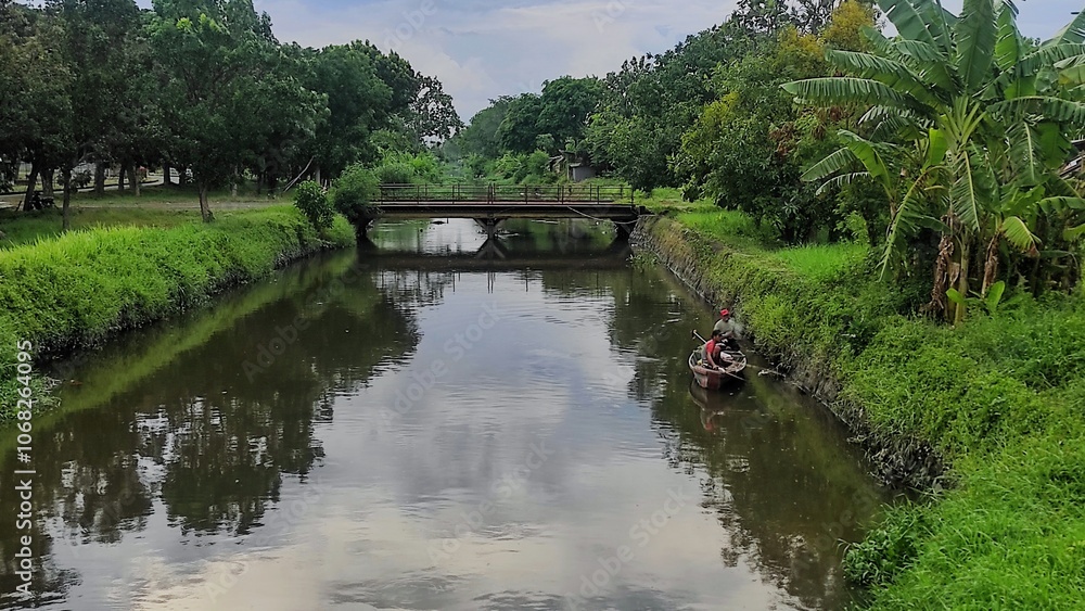 Fototapeta premium A pair of unrecognisable men in a small boat are fishing using electric equipment in the calm river, surrounded by lush greener
