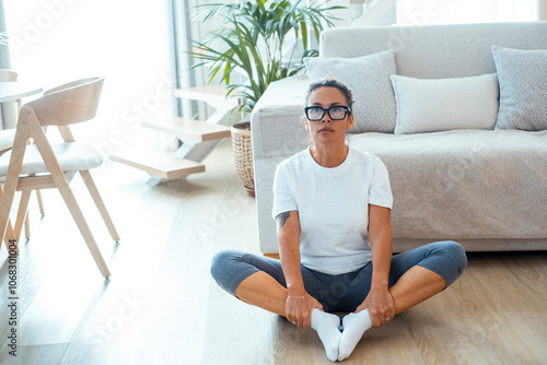 The woman sits on the floor in her apartment, dressed in a white t-shirt and casual sportswear, performing her daily yoga or pilates routine focused on improving her physical and mental wellbeing