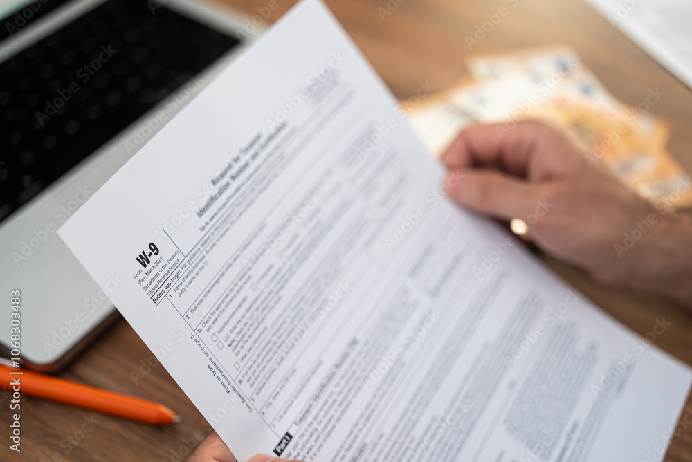 Fototapeta premium Consultant looks over W-9 tax form to provide TIN to client for reporting consulting fees already paid during year at wooden table with euro bills and laptop during break