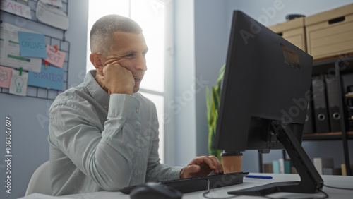 Young man working in an office, sitting at a desk with a computer, looking focused, and wearing a light blue shirt in a bright, modern workplace with organized files and notes on a bulletin board