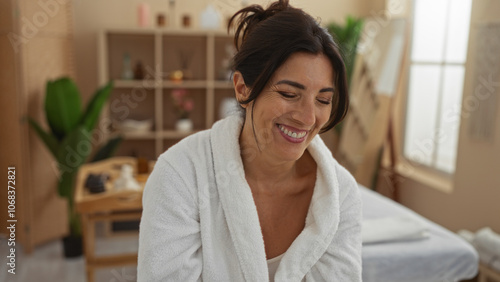 Woman smiling in a spa room, dressed in a white robe, surrounded by a calm, wellness-focused environment with soft lighting and greenery.