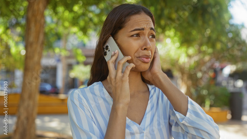 Young woman talking on the phone looks concerned in an urban park setting, with green trees and city buildings in the background on a sunny day.