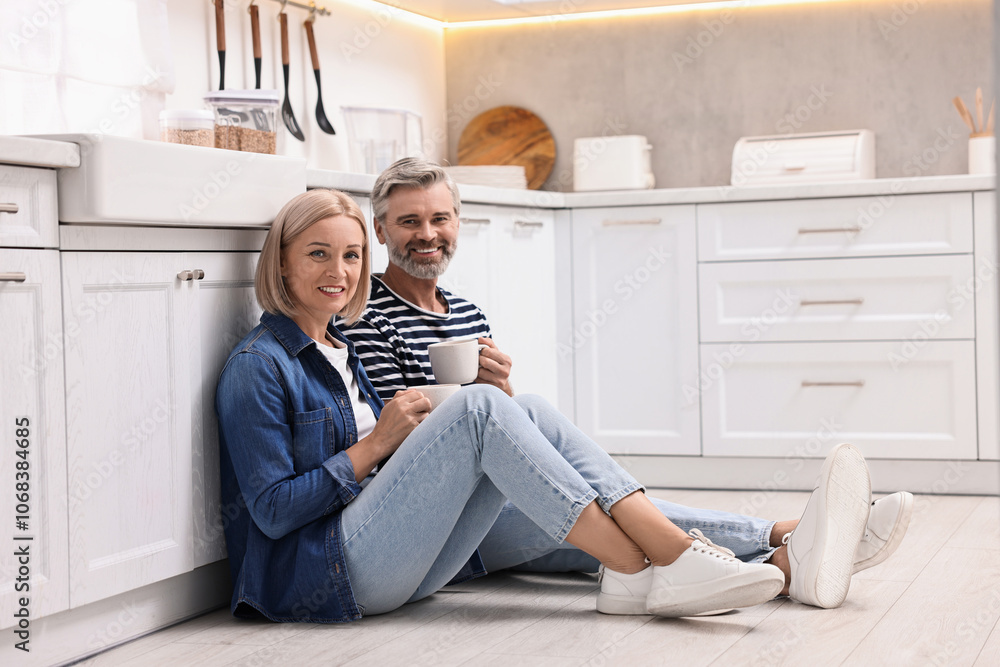 Happy middle aged couple with cups of drink on floor in kitchen Happy middle aged couple with cups of drink on floor in kitchen