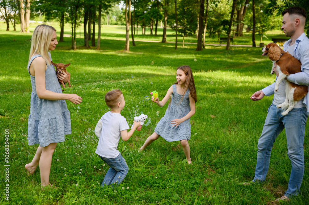 Children playing with soap bubbles in park. Boy and girl blowing soap bubbles from toys, happy parents standing nearby holding pets, dogs. Concept of happy childhood, summer holidays, family weekend Children playing with soap bubbles in park. Boy and girl blowing soap bubbles from toys, happy parents standing nearby holding pets, dogs. Concept of happy childhood, summer holidays, family weekend