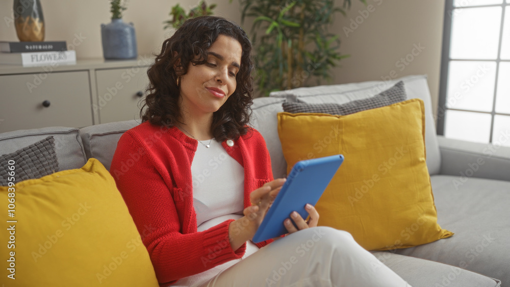 A young attractive hispanic woman with brunette hair enjoys relaxing in her living room, using a tablet while seated on a gray sofa with yellow pillows. A young attractive hispanic woman with brunette hair enjoys relaxing in her living room, using a tablet while seated on a gray sofa with yellow pillows.
