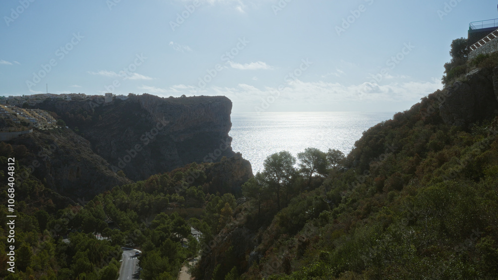 Naklejka premium A scenic coastal landscape featuring cliffs overlooking the mediterranean sea, lush greenery, and distant houses under a clear sky.