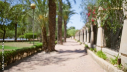 Blurry image of outdoor park pathway with trees, benches, and a fence under bright sunlight