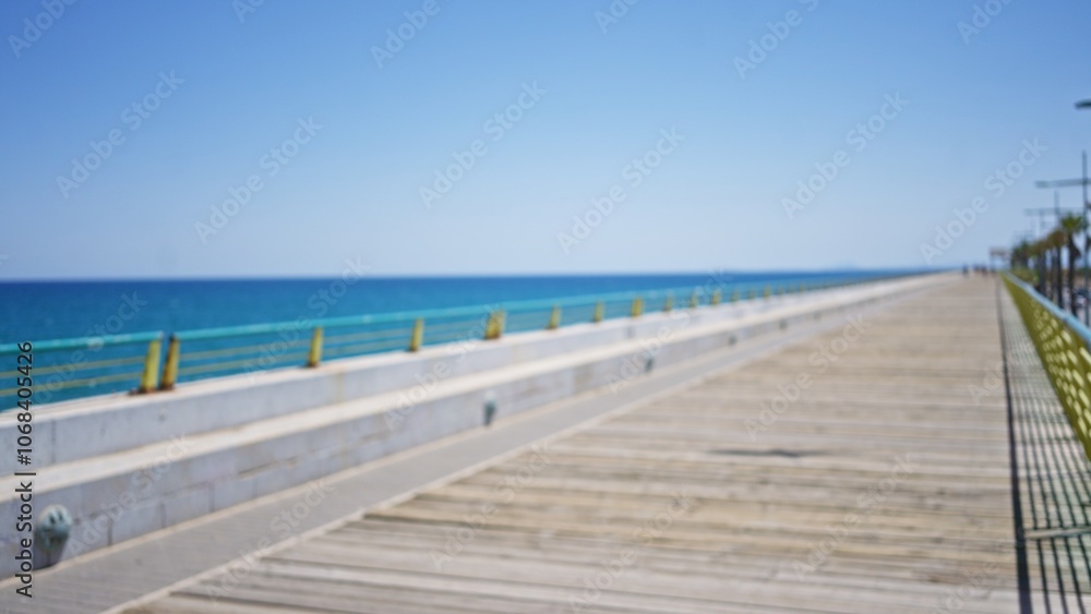 Blurred pier extending into the sea under a clear blue sky with distant defocused figures walking and bokeh effect creating a serene outdoor coastal scene Blurred pier extending into the sea under a clear blue sky with distant defocused figures walking and bokeh effect creating a serene outdoor coastal scene