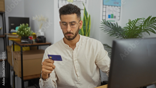 Young hispanic man in an office, looking at a credit card while sitting at a desk with a computer, surrounded by plants and office supplies.