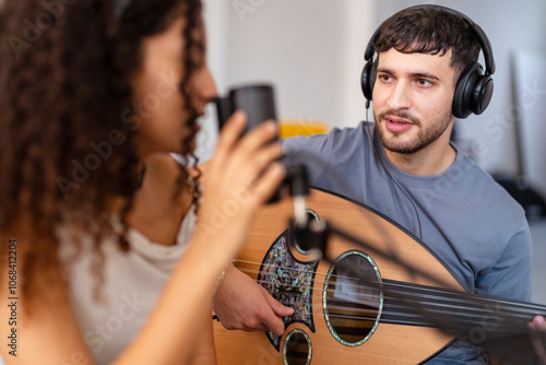 A young man plays an oud while a woman sings into a microphone, both wearing headphones, engaging in a focused recording session in a cozy home setting emphasizing creativity and teamwork. photo