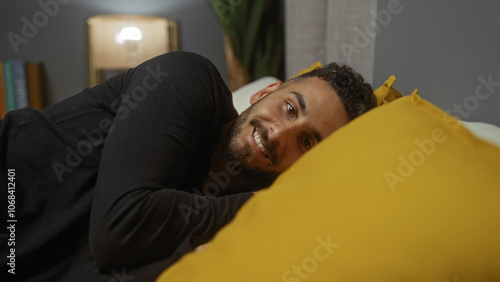 Handsome young hispanic man lying on a bed in a cozy bedroom at home, smiling warmly at the camera with a yellow pillow in the foreground.