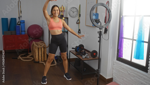 Woman recording fitness video in home gym with exercise equipment, smiling for followers while wearing workout clothes and standing near ring light