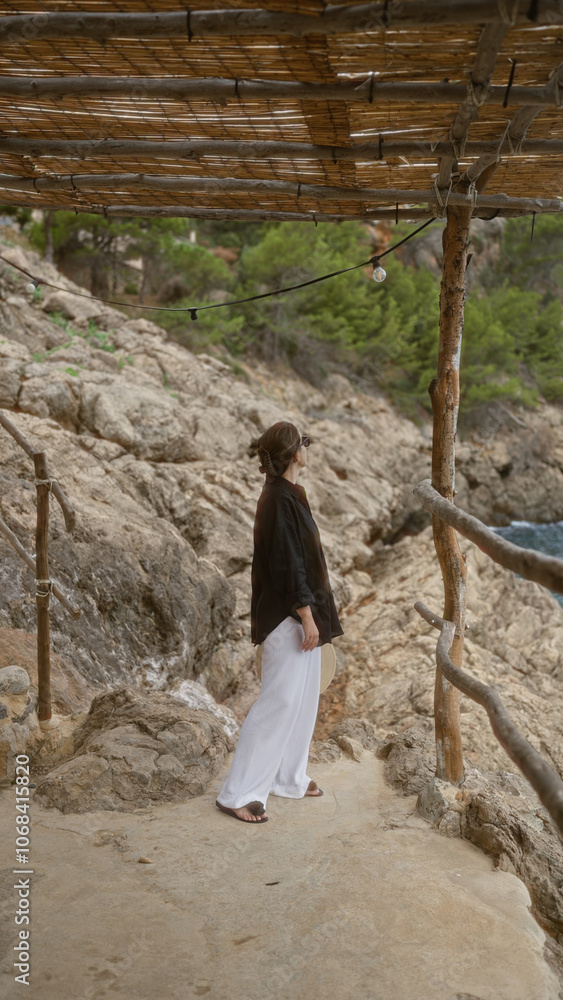 Woman in cala deia, mallorca, stands outdoors by rocky seashore under wooden canopy, wearing white trousers, black shirt, and sunglasses, with lush greenery of the spanish island in the background