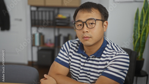 A handsome young chinese man in glasses and a striped shirt sits in an office with bookshelves in the background.