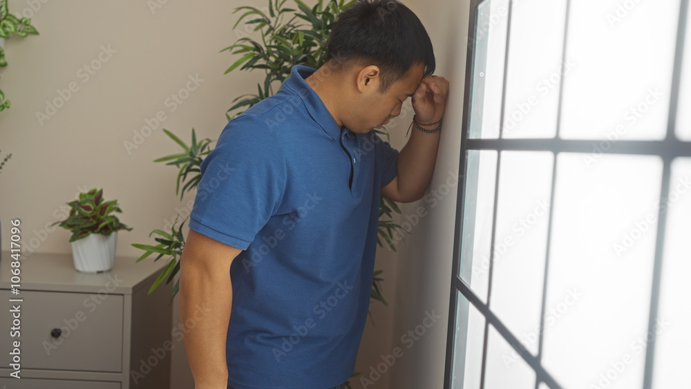 Young chinese man leaning against a wall in a living room with plants, depicting a moment of contemplation. Young chinese man leaning against a wall in a living room with plants, depicting a moment of contemplation.