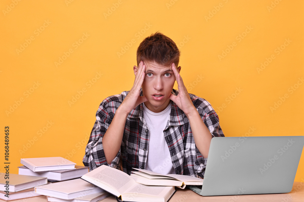 Young student with books and laptop having stress before exam at table against yellow background Young student with books and laptop having stress before exam at table against yellow background