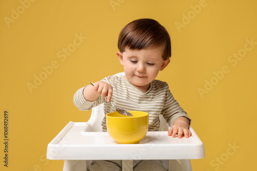 Cute little kid eating healthy baby food from bowl in high chair on yellow background