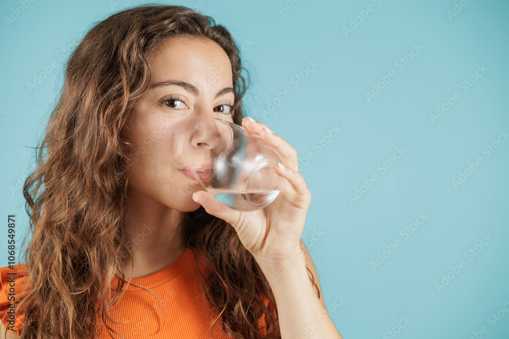 Portrait of young woman on blue background. She holds glass cup in her hand and drinks water. Hydrating herself to be healthy. Woman isolated. Portrait of young woman on blue background. She holds glass cup in her hand and drinks water. Hydrating herself to be healthy. Woman isolated.