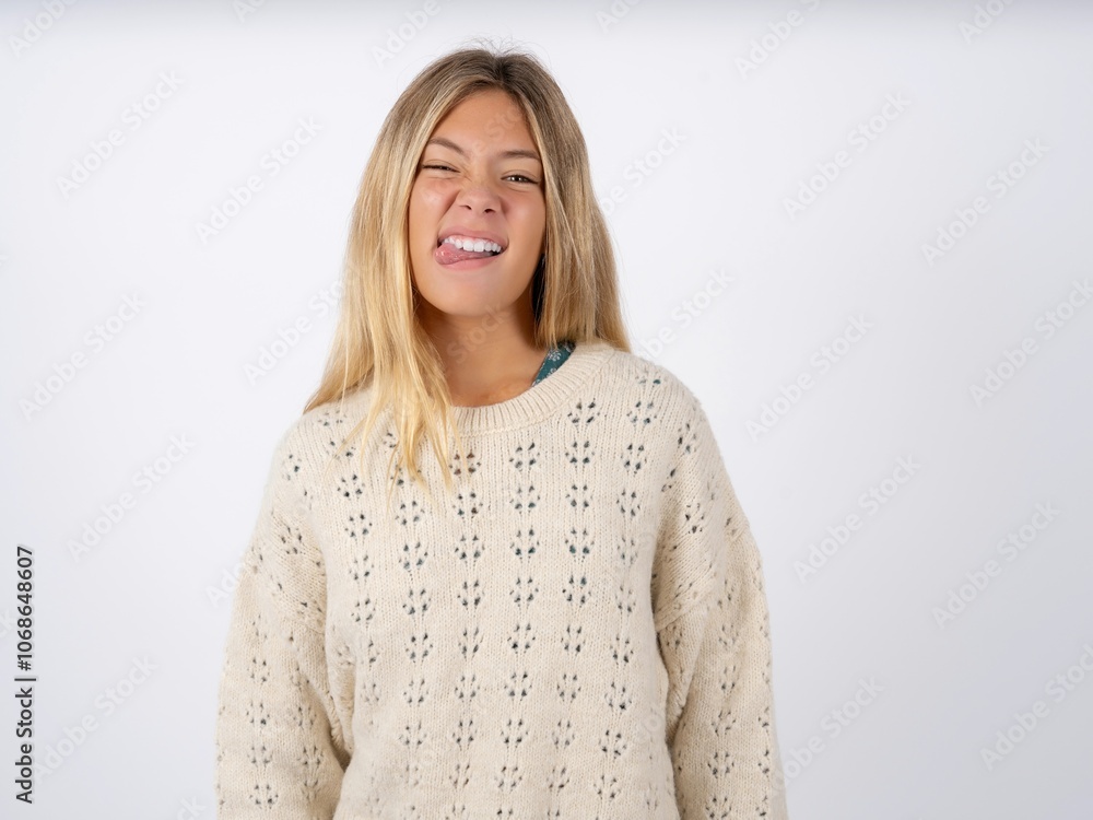 Funny caucasian teen girl wearing knitted sweater over white makes grimace and crosses eyes plays fool has fun alone sticks out tongue. Funny caucasian teen girl wearing knitted sweater over white makes grimace and crosses eyes plays fool has fun alone sticks out tongue.