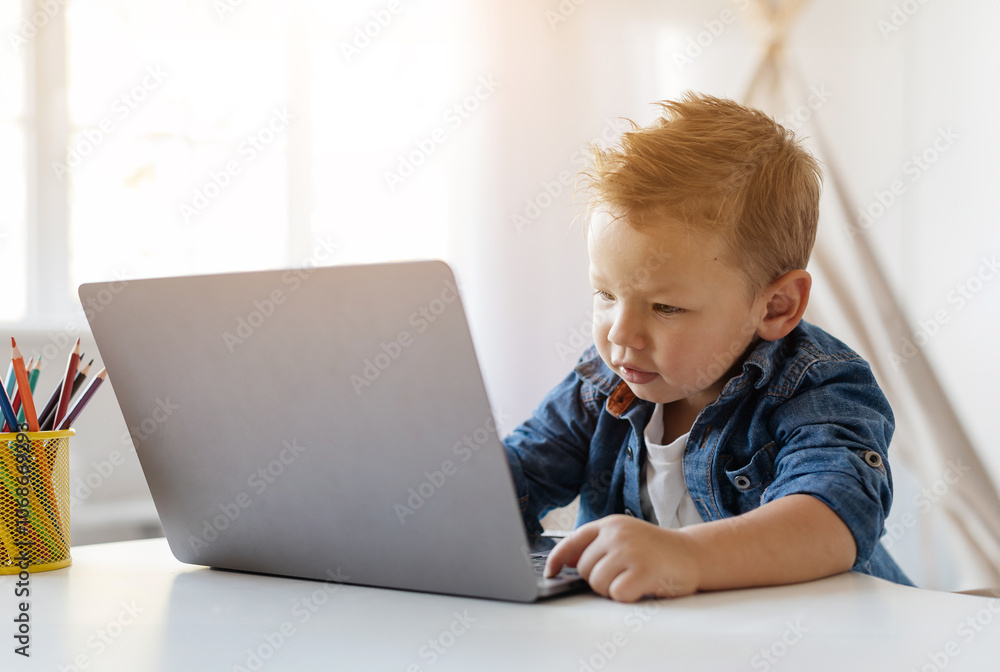 Adorable little toddler boy sits at a table, intently working on a laptop. Sunlight filters through the window, illuminating his concentration and curiosity during a playful afternoon. Adorable little toddler boy sits at a table, intently working on a laptop. Sunlight filters through the window, illuminating his concentration and curiosity during a playful afternoon.