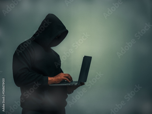 Faceless anonymous hacker in black hoodie and covering his face holding a laptop with a light shining down from above and dark background,Financial data theft concept, Empty space for text, copy space