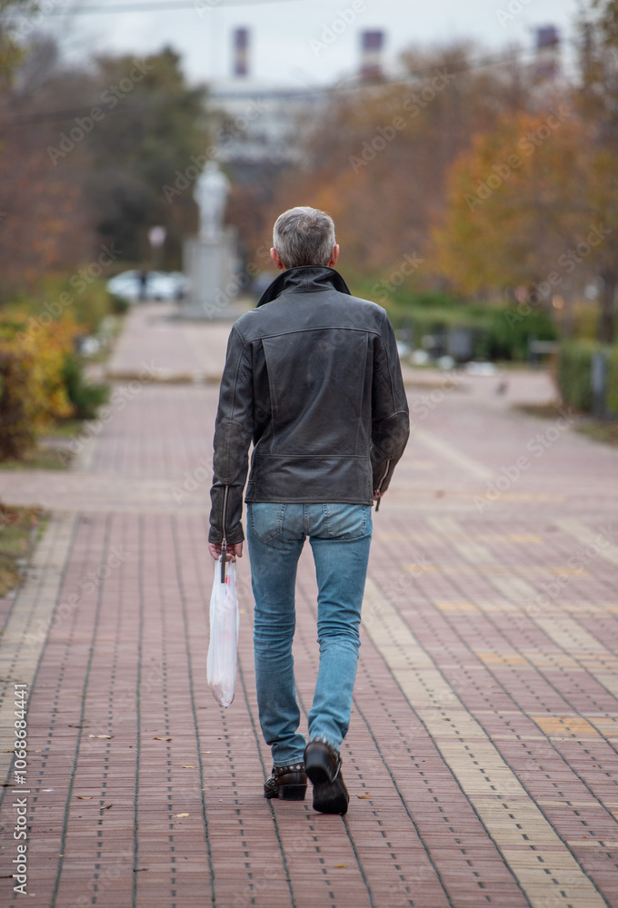 A man wearing a leather jacket and blue jeans walks down a brick walkway A man wearing a leather jacket and blue jeans walks down a brick walkway