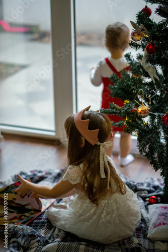 two girls near the Christmas tree, one decorates the tree, the other girl reads a book