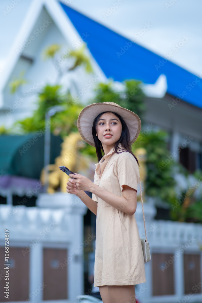 Fototapeta premium A woman in a tan dress is holding a cell phone and wearing a straw hat