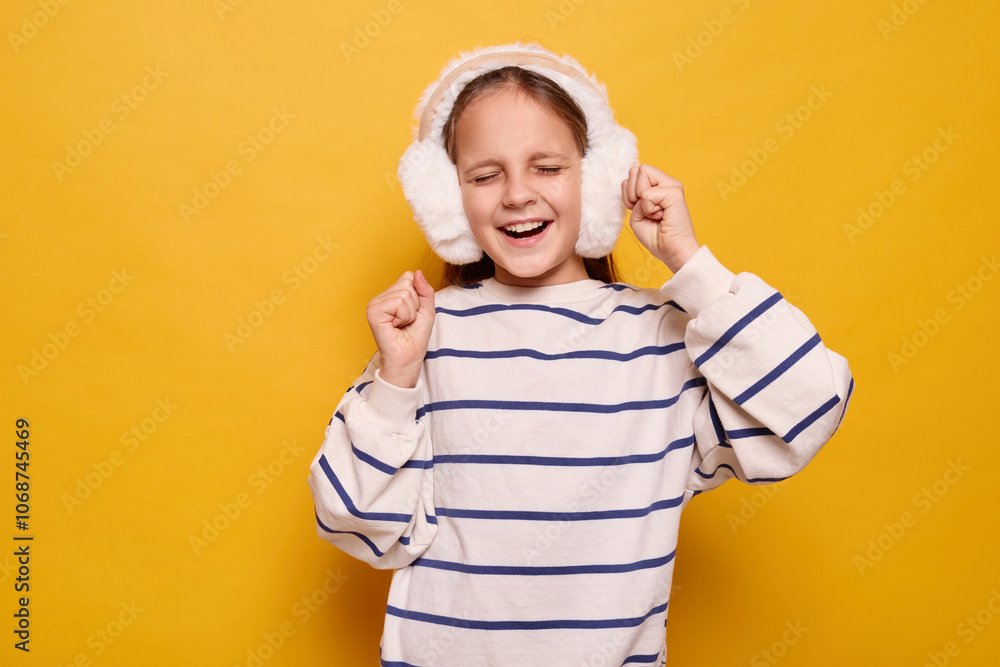 Overjoyed little girl with fur headphones dancing joyfully celebrating triumph and victory feeling excited enjoying moment with happy expression isolated on yellow background Overjoyed little girl with fur headphones dancing joyfully celebrating triumph and victory feeling excited enjoying moment with happy expression isolated on yellow background