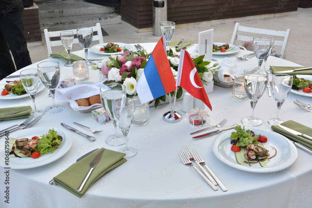 A table set for a formal dinner, featuring Turkish and Russian flags as a centerpiece. The elegant tableware and fresh flowers create a sophisticated and inviting atmosphere
