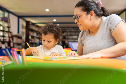Latina teacher smiling as she guides her preschool student in an art project where the child is coloring. A library is visible in the background, copy space.